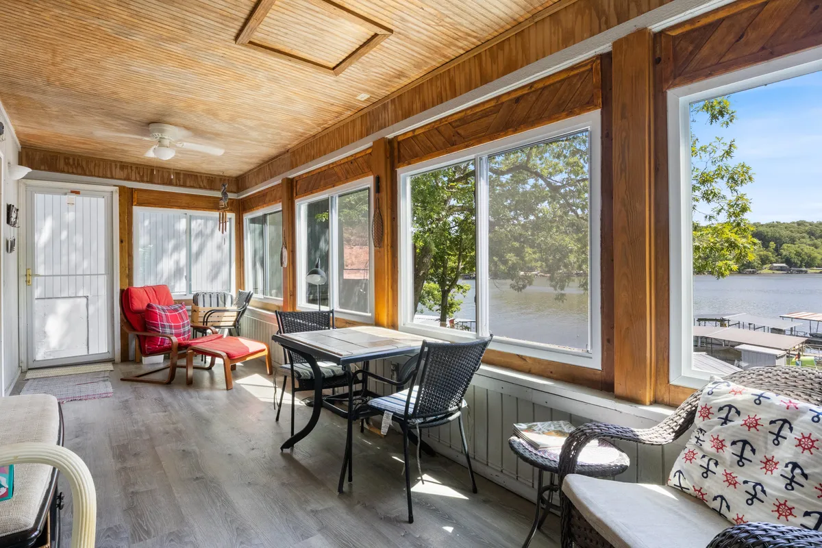 A tranquil, cozy living area featuring comfortable seating grouped around a dining table. Large windows dominate one wall, framing sweeping views of the lake and allowing sunlight to pour in. The arrangement invites both relaxation and social gathering, with the lake’s beauty visible from every angle.