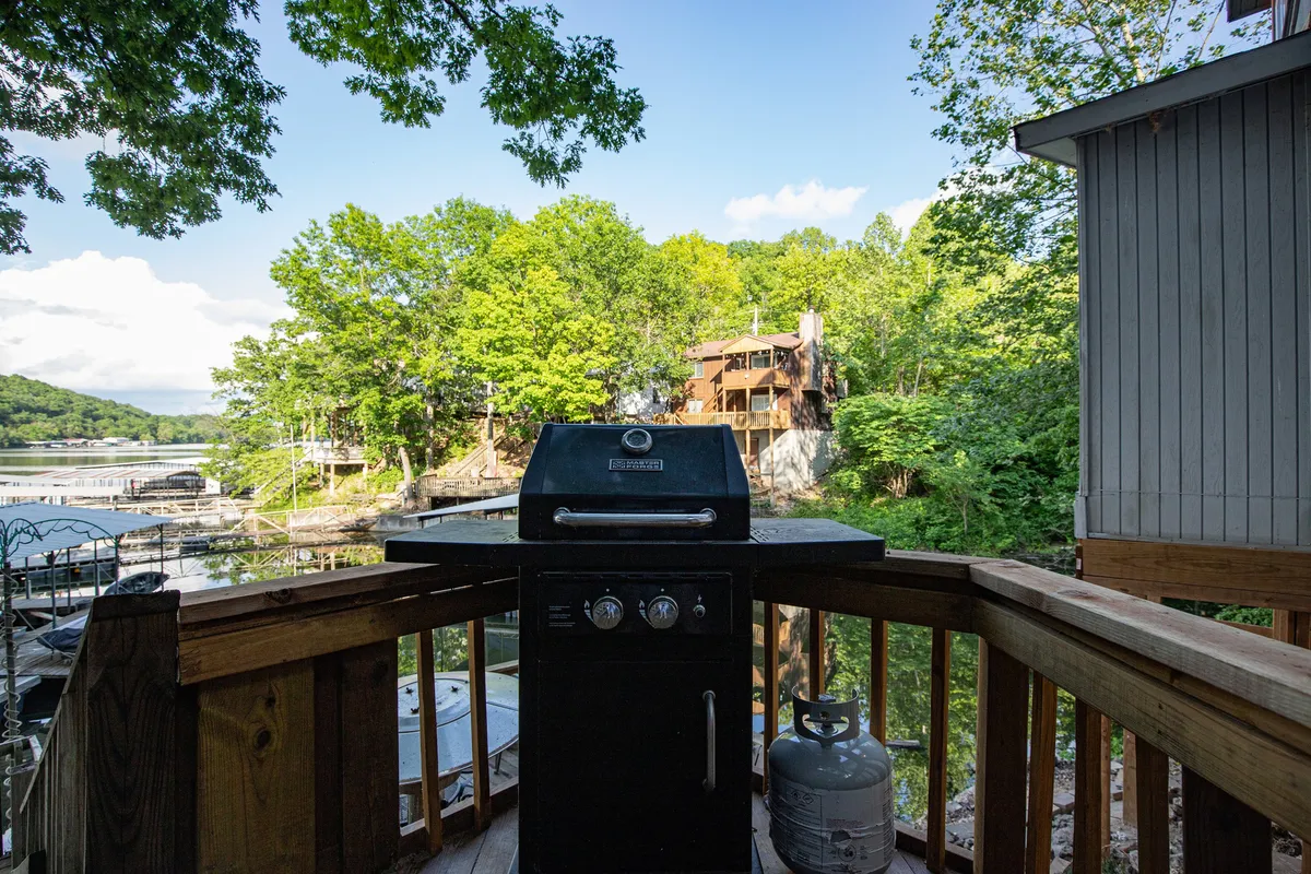 An inviting exterior scene with a grill set up on a deck or patio area, surrounded by lush greenery and with a scenic lake view in the background. The setup suggests a space ideal for outdoor dining and group gatherings, emphasizing a communal and celebratory atmosphere.
