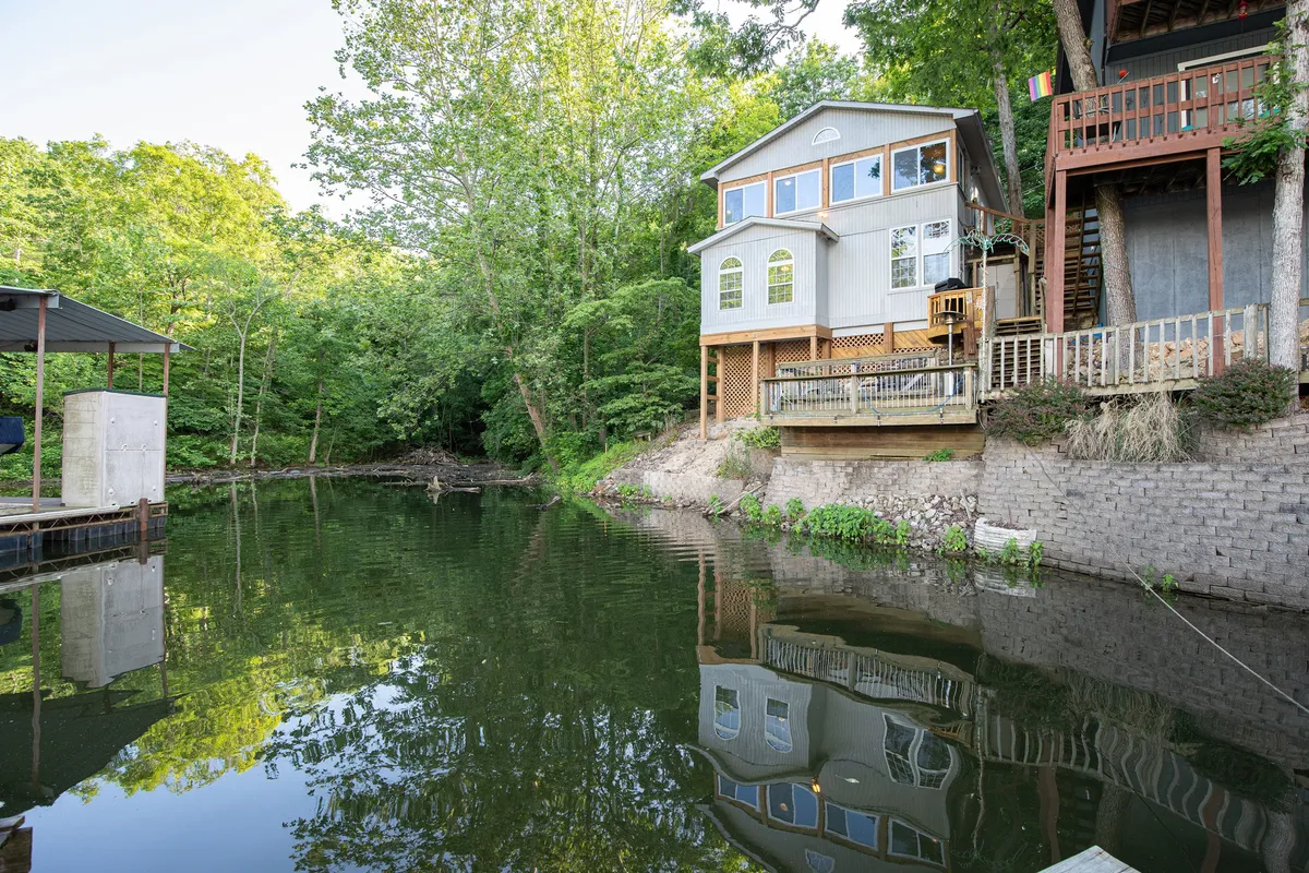 The image showcases a charming cottage nestled among trees, perched on the rocky shore of Lake of the Ozarks. The house overlooks calm, sparkling water, with a spacious deck facing the lake—perfect for relaxing, fishing, or savoring a peaceful sunrise. The scene exudes tranquility, with hints of sunset lighting and the promise of outdoor adventures.