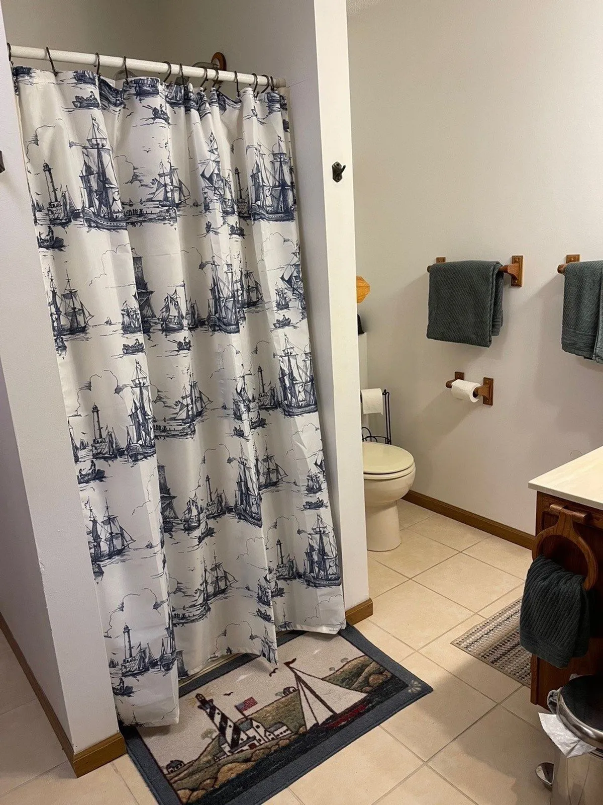 A bathroom with a nautical shower curtain, a lighthouse-themed bath mat, and a towel rack neatly arranged with gray towels. The space is clean, fresh, and inviting, offering both functionality and a touch of fun nautical decor.