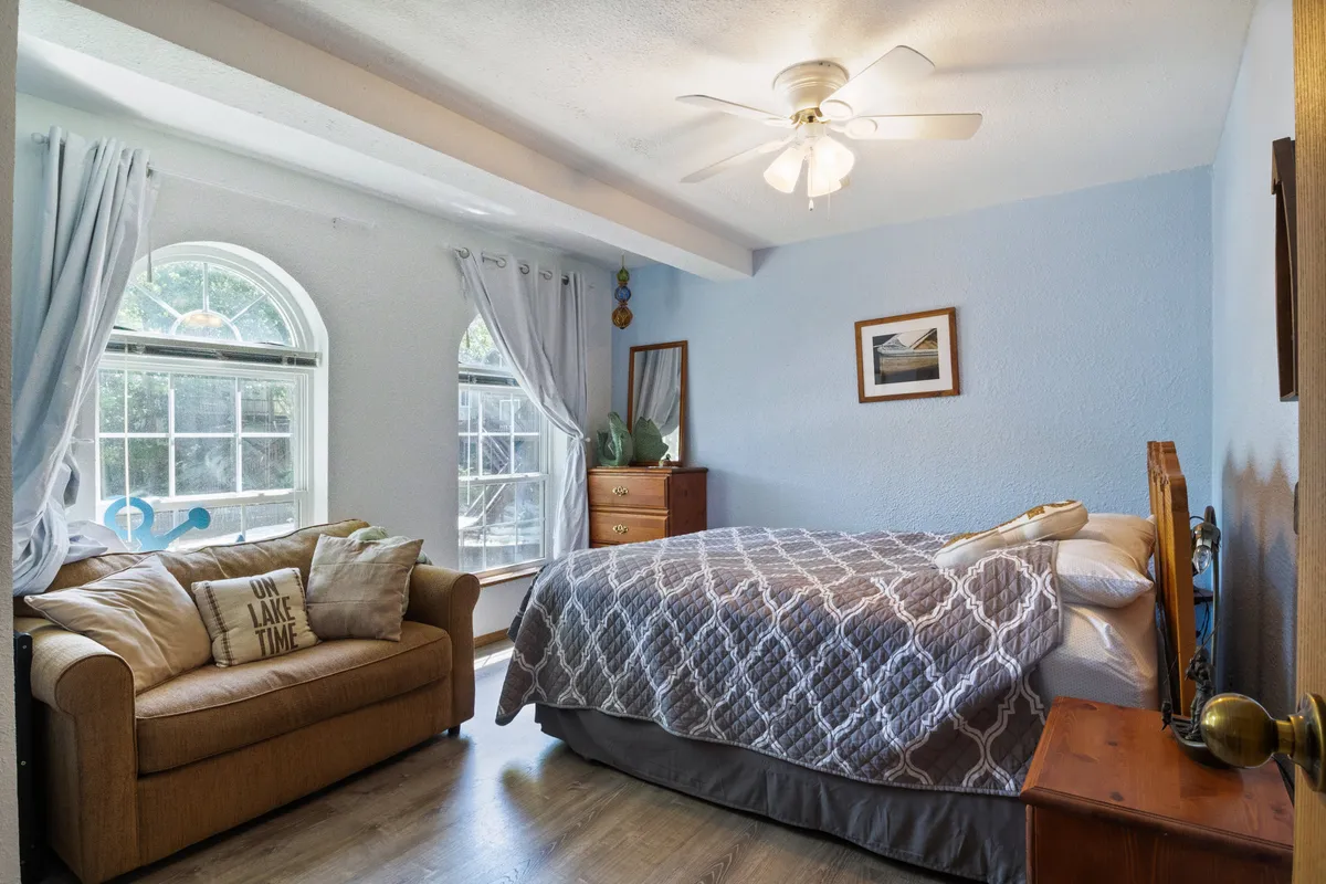 This image shows a bright bedroom with a bed featuring a gray quilt, a cozy sofa adorned with decorative pillows, a wooden dresser, and a large window letting in abundant natural light. The artwork and overall styling create a serene, comfortable atmosphere ideal for relaxing or reading.