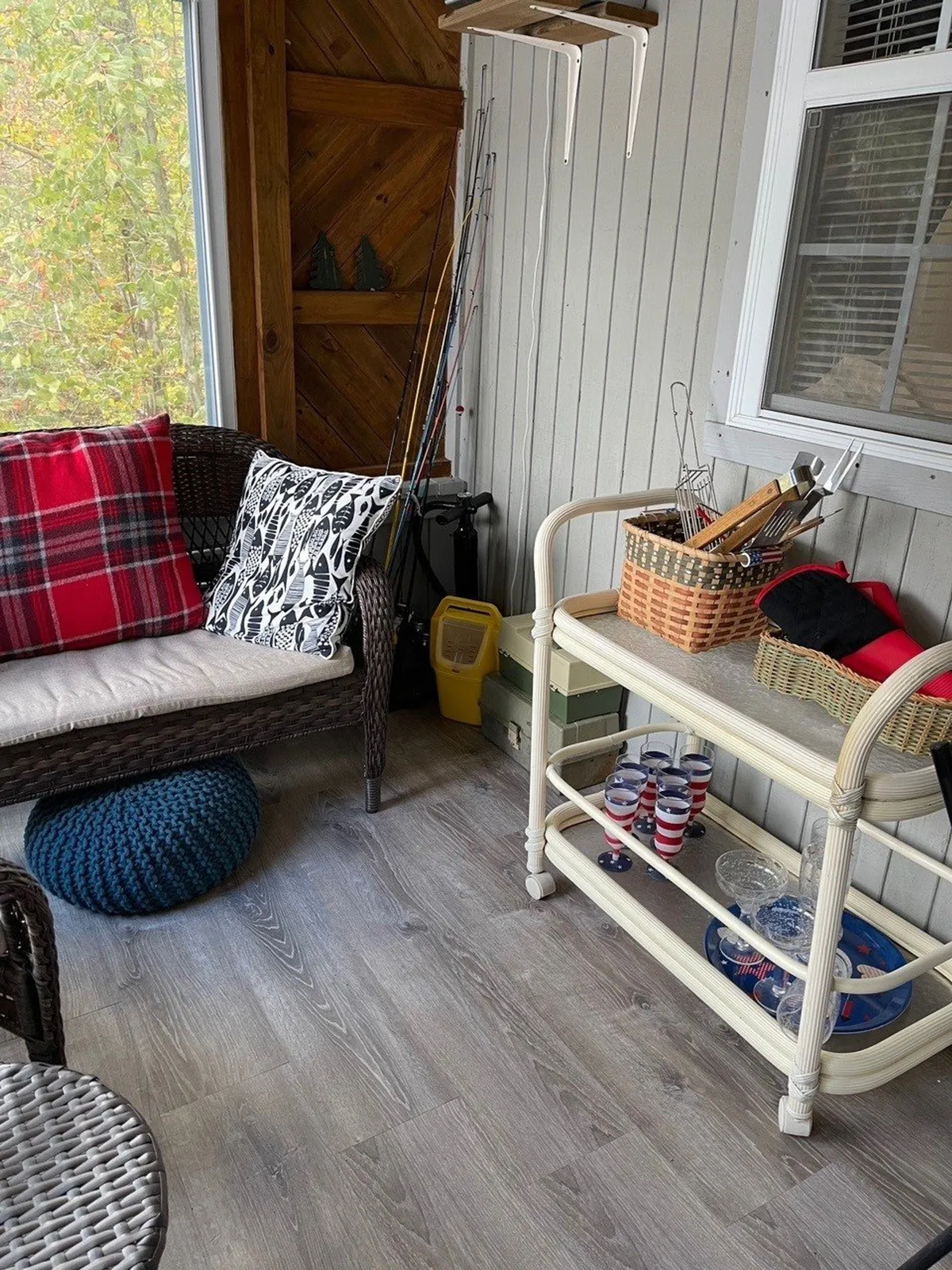 A cozy sun porch furnished with wicker and cushioned seating, plaid and decorative pillows, and a shelving unit filled with kitchen items and fishing rods. Large windows on multiple sides allow for expansive lake and nature views, and the welcoming decor invites long, leisurely conversations.