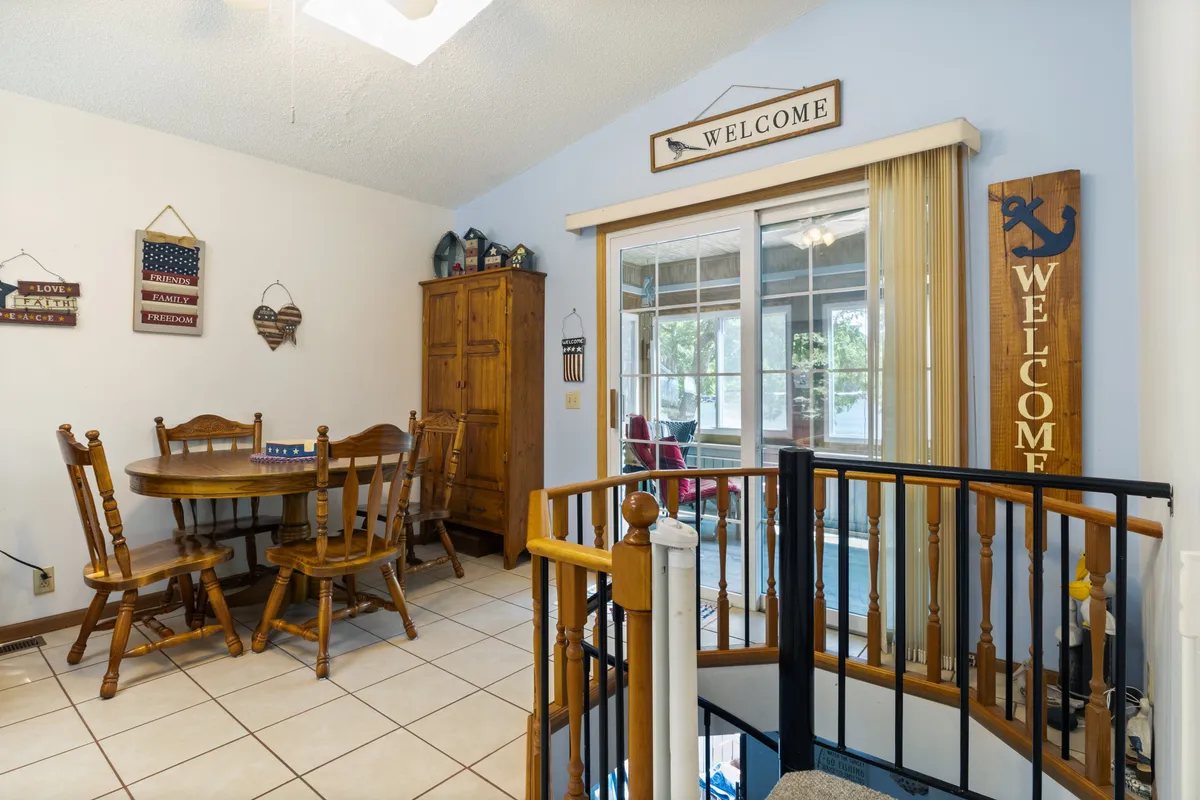 A warm, welcoming dining area with a wooden table and chairs, wooden cabinet, and playful welcome signs. Large sliding doors lead to a sunroom, hinting at the seamless flow between indoor and sun porch spaces. The spiral staircase adds character and a sense of homey charm.