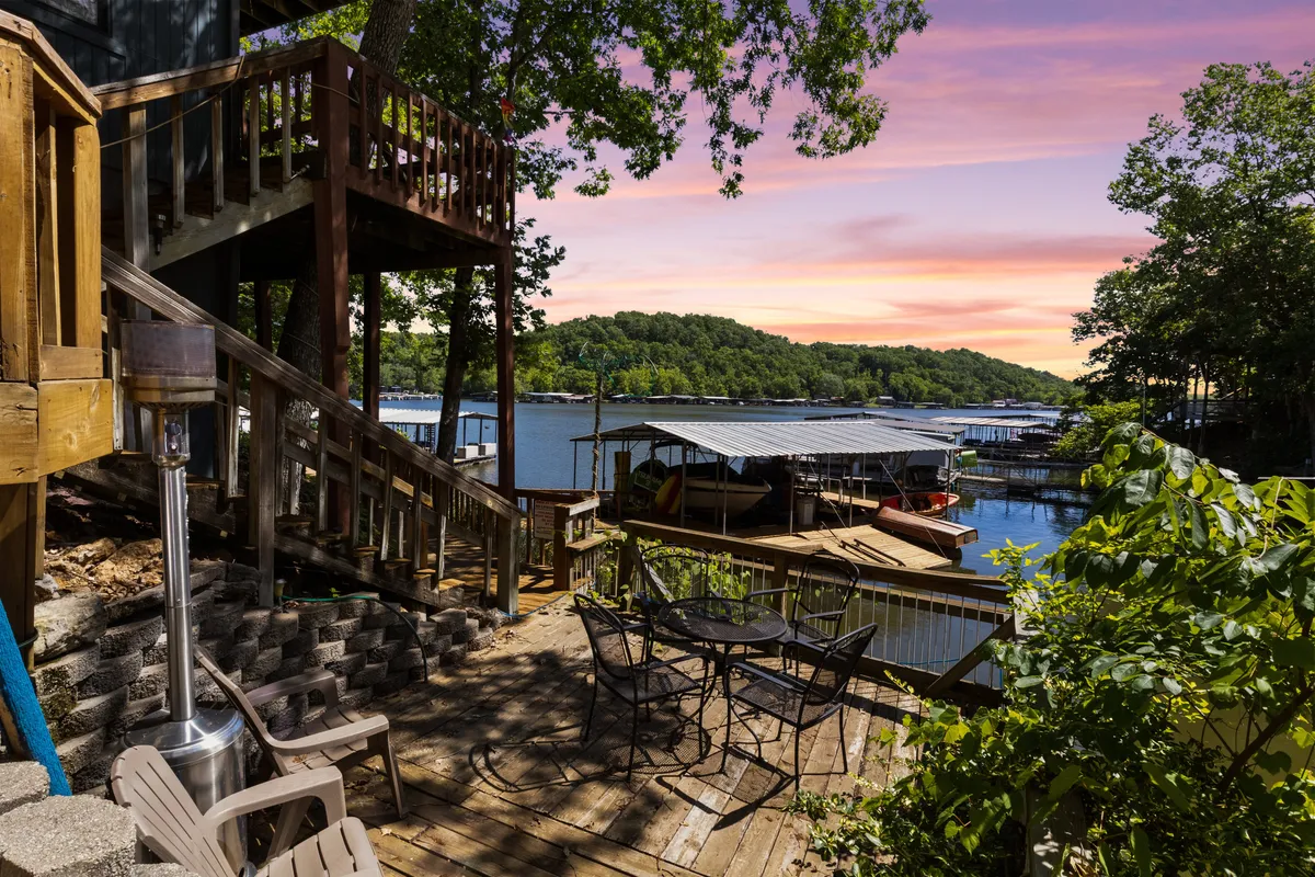 An exterior deck with wooden seating, offering stunning views of the lake, a staircase leading up, and boathouses in the background. The space is ideal for outdoor dining, relaxing with friends, or enjoying morning coffee with a breeze and lake views.