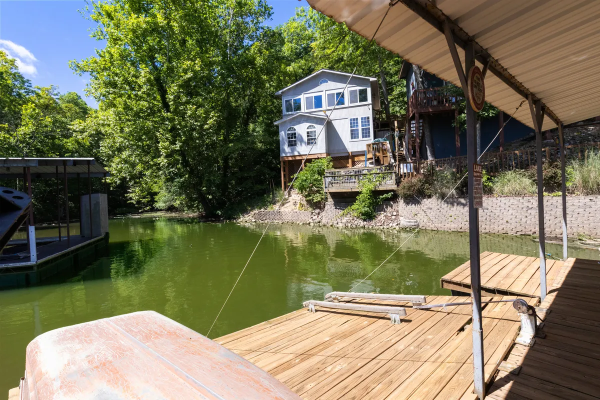A scenic view of the waterfront dock with the house featuring large windows set amidst surrounding greenery. The dock extends into calm water, inviting for fishing, swimming, or launching a kayak. The ample outdoor space hints at opportunities for barbecues and lakeside gatherings.